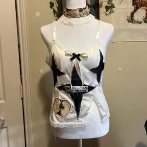 May include: White satin camisole with black star-shaped patches and a small black bow. The camisole features a lace trim and a half-apple patch. A matching choker with floral accents completes the look. The camisole is displayed on a mannequin.