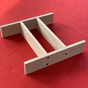 May include: A light-colored wooden shelf with three sections. The shelf is rectangular with two vertical supports and two horizontal supports. The wood has a natural grain and is set against a red background.