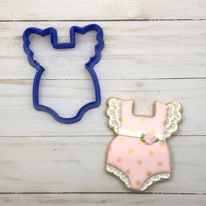 May include: A blue plastic cookie cutter in the shape of a baby onesie sits next to a decorated cookie. The cookie is pink with gold polka dots, white trim, and a small pink rose. The cookie is designed to look like a baby onesie.