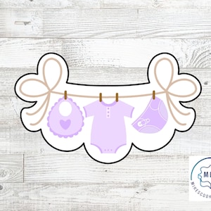 May include: A purple baby onesie, bib, and nappy are hanging on a clothesline with a brown bow at each end. The image is for a cookie cutter set.