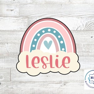 Może przedstawiać: Wycinarka do ciasteczek w kształcie tęczy w kolorze różowym, niebieskim i białym z chmurką i imieniem "Leslie" w różowych literach. Wycinarka do ciasteczek znajduje się na białym drewnianym tle.