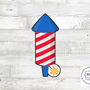 May include: A cartoon-style firework with a blue top and red and white striped body. A sparkler is lit at the bottom. The image is on a white wood grain background. The firework is a cookie cutter.