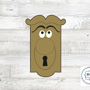 Può includere: Un tagliapasta a forma di pomo di porta in legno marrone con un viso, sopracciglia, occhi e un buco della serratura. Il tagliapasta è progettato per creare un biscotto che assomiglia a un pomo di porta.