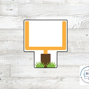 May include: A cookie cutter in the shape of a football goal post. The goal post is yellow and the uprights are black. The goal post is sitting on a green grassy field.