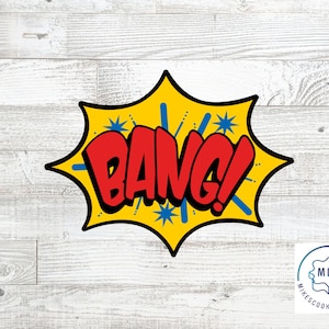 May include: A yellow and blue comic book style speech bubble with the word "BANG!" in red letters. The speech bubble is on a white wood background.