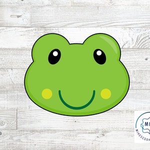 Op de afbeelding: Een groene koekjesvorm in de vorm van een kikkergezicht met zwarte ogen, een lachende mond en gele wangvlekken. De koekjesvorm is zwart omlijnd. De achtergrond is een licht houtnerfpatroon.