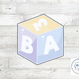 May include: A blue, yellow, and purple 3D block cookie cutter with the letters "B", "A", and "3" on each side.  The cookie cutter is on a white wood background.
