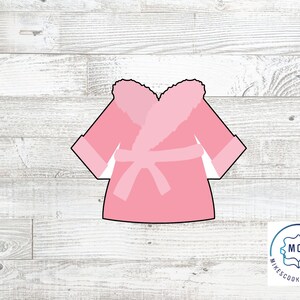 May include: Pink bathrobe cookie cutter with a fluffy collar and a sash. The cutter is on a white wood background.