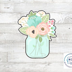 May include: A cookie cutter in the shape of a mason jar filled with flowers. The flowers are pink, blue, and white. The jar is light blue. The cookie cutter is made by Mike's Cookie Cutters.