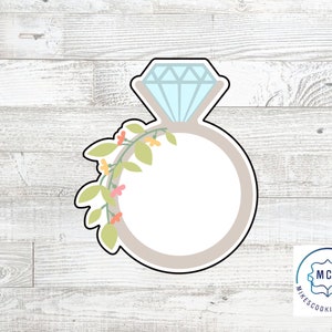 May include: A cookie cutter in the shape of a ring with a diamond on top. The ring is surrounded by a vine with small flowers. The cookie cutter is made of metal and is light blue, gray, and green.