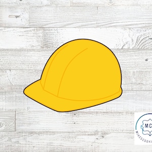 May include: A yellow construction worker hard hat cookie cutter. The cookie cutter is on a white wood background.