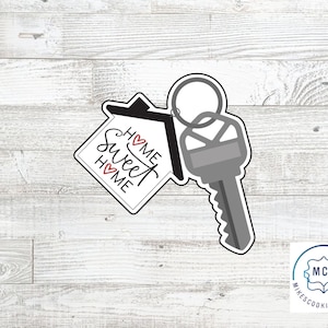 May include: A black and white cookie cutter in the shape of a house with a key attached. The house has the words "Home Sweet Home" written on it with red hearts.