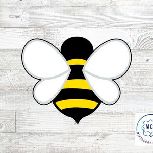 May include: A black and yellow bee cookie cutter with white wings. The cookie cutter is on a white wood background.