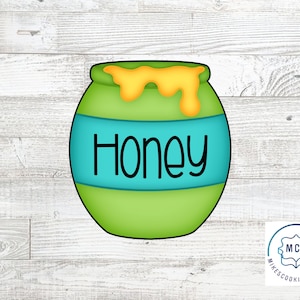 May include: Green honey pot cookie cutter with a blue stripe and the word "Honey" written on it. The honey pot has a yellow drip of honey coming out of the top.