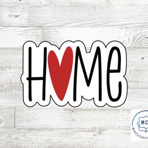 以下が含まれることがあります： 白いクッキーカッターに黒い文字で「HOME」と書いてあり、中央に赤いハートがあります。