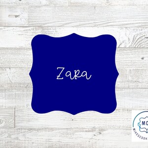 May include: A blue, custom cookie cutter in the shape of a decorative frame with the name "Zara" written in white inside.