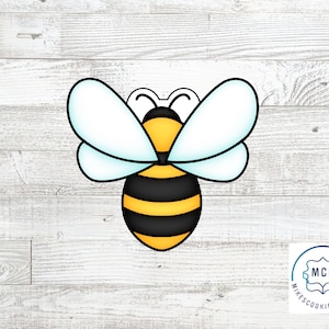 May include: A yellow and black bee cookie cutter with white wings. The cookie cutter is on a white wood background.