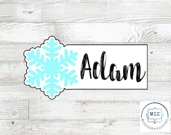 Puede incluir: Un cortador de galletas blanco con un diseño de copo de nieve azul y el nombre "Adam" en letra negra.