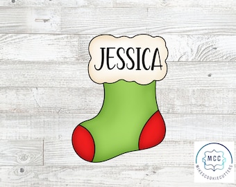 May include: A green and red Christmas stocking cookie cutter with the name "Jessica" on the top. The cookie cutter is on a white wood background.