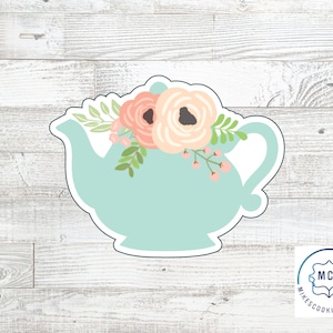 Op de afbeelding: Een lichtblauwe koekjesvorm in de vorm van een theepot, versierd met perzik- en roze bloemen en groene bladeren. De koekjesvorm heeft een handvat en een tuit. De afbeelding staat op een wit en grijs houtnerf-achtergrond.