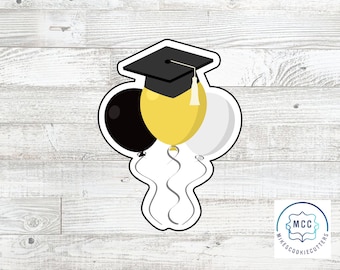 May include: A graduation cookie cutter with a black graduation cap and three balloons, one black, one yellow, and one white.