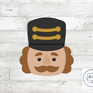 Pode incluir: Um cortador de biscoitos em forma de rosto de quebra-nozes. O quebra-nozes tem um chapéu preto com detalhes dourados, um rosto bronzeado, cabelo castanho e um bigode castanho. O cortador de biscoitos está em um fundo de grão de madeira clara.