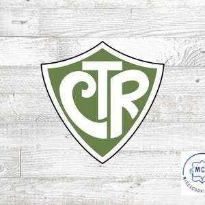 Könnte beinhalten: Grüner schildförmiger Ausstecher mit den Buchstaben "CTR" in Weiß darin. Der Ausstecher befindet sich auf einem weißen Holzgrund.