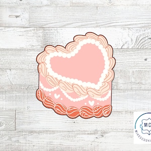 May include: Pink heart-shaped cake cookie cutter with white frosting and red accents.  The cutter is on a white wood background.  Mike's Cookie Cutters logo is in the lower right corner.