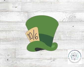 May include: A green top hat cookie cutter with a dark green band and a tan rectangular tag attached to the side. The tag features the black text 10/6 printed clearly on its surface. The design is flat and features a wide brim and tall crown.