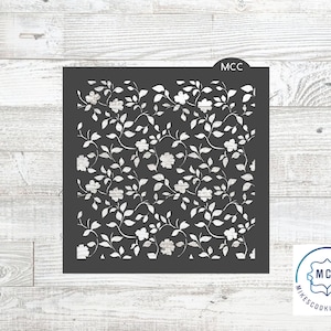 May include: A square, dark gray stencil featuring a floral pattern of white flowers and leaves. The stencil is designed for crafting and decorating. The text "MCC" is visible at the top of the stencil.