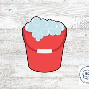 May include: Red plastic bucket filled with white foam. The bucket has a white handle and is on a white wood background.
