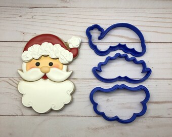 Puede incluir: Una galleta de Papá Noel y tres cortadores de galletas de plástico azul. La galleta tiene un gorro rojo, barba blanca y una cara sonriente. Los cortadores de galletas tienen forma de gorro, barba y cara de Papá Noel.