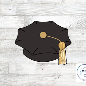Peut inclure: Un chapeau de graduation noir avec un gland et un bouton dorés. La forme du chapeau est celle d'un emporte-pièce, parfait pour les créations sur le thème de la remise de diplômes. Fond en bois clair.