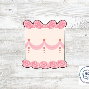 May include: Pink and white cookie cutter shaped like a cake with pink frosting and a pink ribbon design. The cutter is on a white wood background.