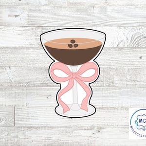 May include: A cookie cutter in the shape of a cocktail glass filled with a brown liquid and three coffee beans. A pink bow is tied around the stem of the glass. The cookie cutter is white with a black outline.