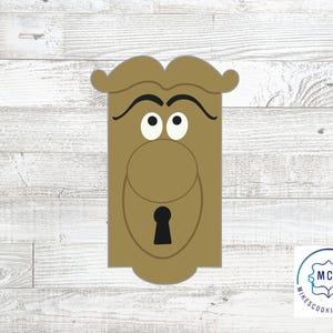 May include: A tan cookie cutter shaped like a door knob with a face, featuring two black arched eyebrows, two white eyes with black pupils looking upward, a round central knob, and a black keyhole shape at the bottom. The logo MCC is visible.