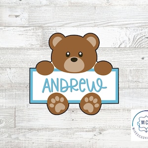 Può includere: Un tagliapasta a forma di orso marrone che tiene un cartello blu e bianco con il nome "Andrew" scritto in corsivo.