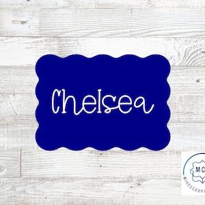 May include: A dark blue cookie cutter with a scalloped edge. The word "Chelsea" is written in white cursive script across the center. The background is a light wood grain pattern.