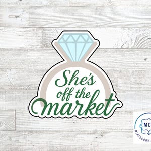 Może przedstawiać: Wykrawacz do ciastek w kształcie pierścionka zaręczynowego z diamentem. Pierścionek jest jasnoszary z jasnoniebieskim diamentem. Tekst "She's off the market" jest w kolorze zielonym.