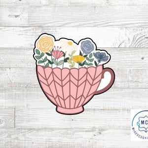 May include: A pink cookie cutter in the shape of a teacup filled with colorful flowers. The flowers include yellow roses, white daisies, pink blooms, and blue flowers. The teacup has a geometric pattern.