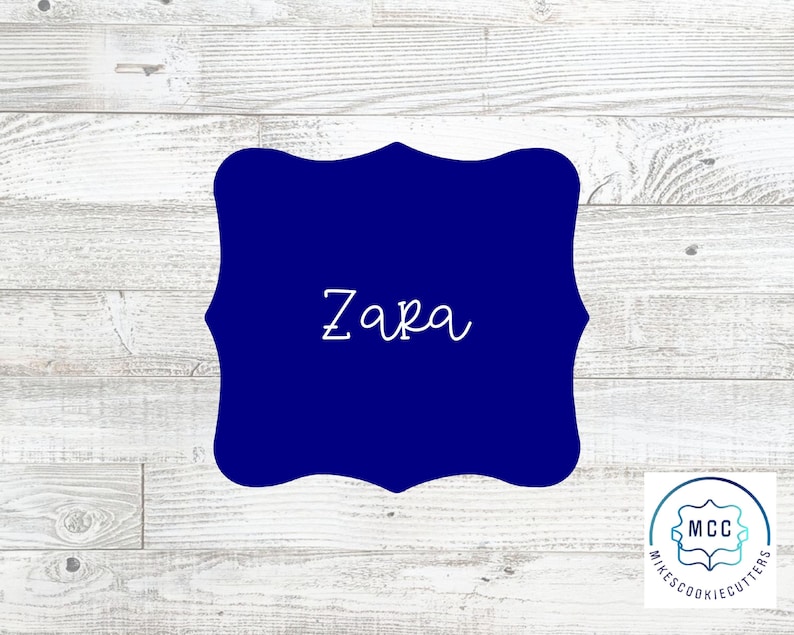 May include: A blue, custom cookie cutter in the shape of a decorative frame with the name "Zara" written in white inside.