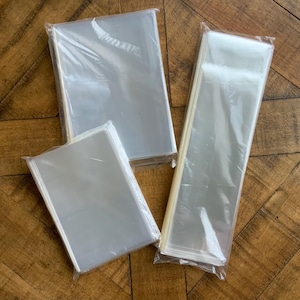 May include: Three clear, sealed plastic bags of varying sizes are arranged on a wooden surface. The bags appear to be empty and are transparent, allowing a view of the contents within. The bags are rectangular and sealed.