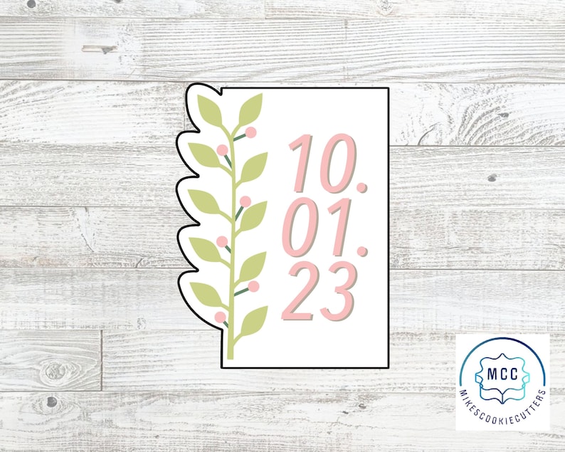 May include: A pink and green cookie cutter with a leafy design and the date 10.01.23. The cookie cutter is on a white wood background.