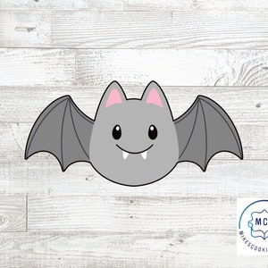 May include: A gray bat cookie cutter with pink ears and a toothy grin. The cutter is on a white wood background.