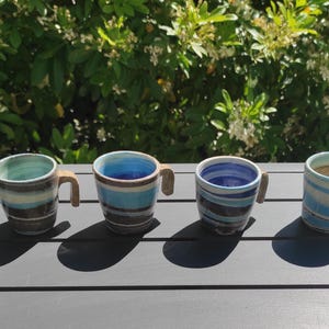 Peut inclure: Six tasses en céramique faites à la main avec des motifs rayés bleus, turquoise et marron. Chaque tasse a une petite anse incurvée et des motifs uniques. Les tasses sont disposées sur une surface gris foncé avec un fond vert naturel.