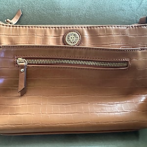May include: A tan crossbody bag with a textured, crocodile-like pattern. The bag has a gold zipper and a small gold emblem. It has a long strap for wearing across the body.
