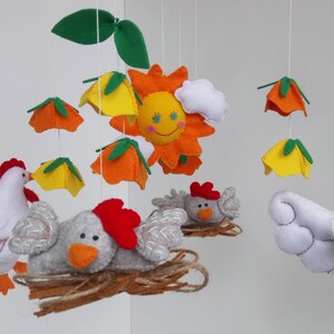 K&ouml;nnte beinhalten: Ein wei&szlig;es, graues und orangefarbenes Filz-Babymobil mit einer Sonne, Bl&auml;ttern und H&uuml;hnern. Die Sonne hat ein l&auml;chelndes Gesicht und eine wei&szlig;e Wolke. Die H&uuml;hner h&auml;ngen mit einer Schnur am Mobile.