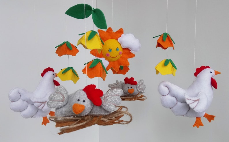 K&ouml;nnte beinhalten: Ein wei&szlig;es, graues und orangefarbenes Filz-Babymobil mit einer Sonne, Bl&auml;ttern und H&uuml;hnern. Die Sonne hat ein l&auml;chelndes Gesicht und eine wei&szlig;e Wolke. Die H&uuml;hner h&auml;ngen mit einer Schnur am Mobile.
