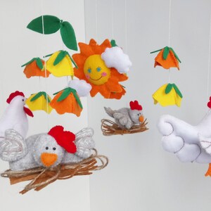 K&ouml;nnte beinhalten: Ein Filz-Babymobil mit einer gelben Sonne, wei&szlig;en Wolken, orangefarbenen und gelben Blumen und zwei wei&szlig;en H&uuml;hnern mit roten K&auml;mmen. Ein Huhn sitzt in einem braunen Nest mit einem grauen K&uuml;ken.