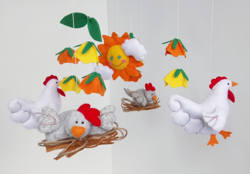 K&ouml;nnte beinhalten: Ein Filz-Babymobil mit einer gelben Sonne, wei&szlig;en Wolken, orangefarbenen und gelben Blumen und zwei wei&szlig;en H&uuml;hnern mit roten K&auml;mmen. Ein Huhn sitzt in einem braunen Nest mit einem grauen K&uuml;ken.